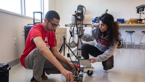 Male student with eyeglasses crounching down and examing infrastructure equipment in an indoor lab with the help of a female student.