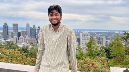 Male college student poses outdoors on a patio overlooking a city skyline. He is smiling wearing a light colored buttoned shirt and jeans.