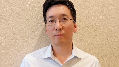 Headshot of man with black hair and glasses wearing an open collared white shirt.
