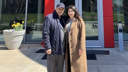 Woman and man pose outside a college building with a red door. The man on the left is wearing a blue baseball cap and blue jacket. The woman has long brown hair and is wearing a camel coat.