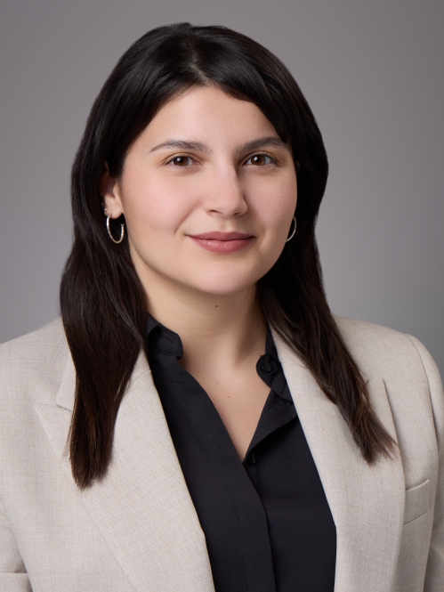 Headshot of woman with long brown hair wearing a black blouse and a light brown jacket.