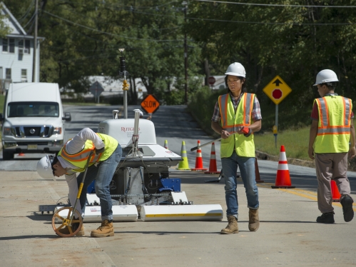 Three men wearing hard hats and yellow safety vests inspecting road with fully automated robotic device for making comprehensive condition assestment. 