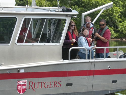 Group of four students standing in the bow of a boat in a river. The boat has the Rutgers logo on the side.
