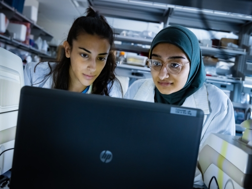 Two female graduate students are looking at a computer screen. The woman on the left has long brown hair. The woman on the right has glasses and is wearing a green headscarf. They both have on white lab coats.