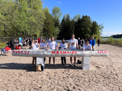 Civil engineering students pose with their concrete canoe on the sandy banks of a lake.