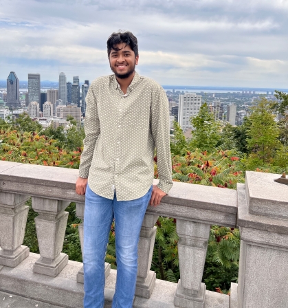 Male college student poses outdoors on a patio overlooking a city skyline. He is smiling wearing a light colored buttoned shirt and jeans.