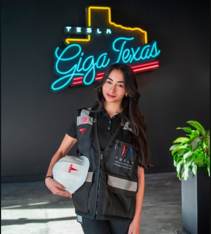 Female engineering student poses wearing a work vest and holding under her arm a hard hat. She is standing in front of the neon sign that says Giga Texas. She has long black hair.