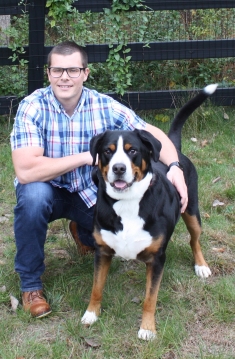 man with eyeglasses wearing a plaid shirt and jeans down on one knee with his arm around a medium size dog next to him